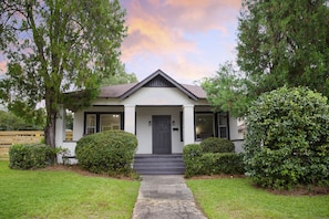 Uma casa de dois andares com varanda frontal, porta escura e pequeno jardim com um caminho que leva à entrada.