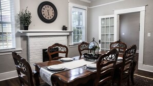 Comedor con una mesa de madera dispuesta para seis comensales, rodeada por seis sillas. Se observa un reloj de pared grande y una planta en maceta.
