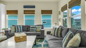 A living room with two grey sofas, a coffee table, and a wicker basket. There are three windows with blue shutters and a sign above them that reads 'Beachfront.' - Beach Bungalow With Private Hot Tub (Bolivar Peninsula)