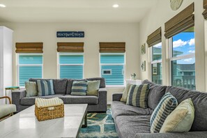 A living room with two grey sofas, a coffee table, and a wicker basket. There are three windows with blue shutters and a sign above them that reads 'Beachfront.'
