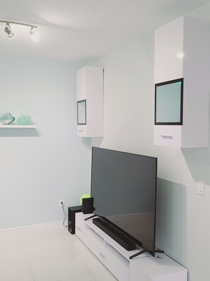 A modern, minimalist room with a large flat-screen TV mounted on a white cabinet, a small shelf with decorative items, and a clean, white tiled floor.