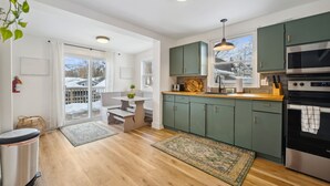 A modern kitchen with green cabinets, a wooden countertop, and stainless steel appliances. There is a dining area with a table and chairs, and a view of a snowy backyard through large windows. - Welcome to The Palms! Your summer retreat ~Hot tub (Fort Erie)