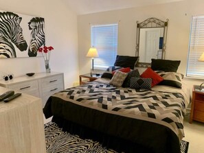 Une chambre à coucher comprenant un lit, une table de chevet, une commode, un miroir et une fenêtre avec des stores.