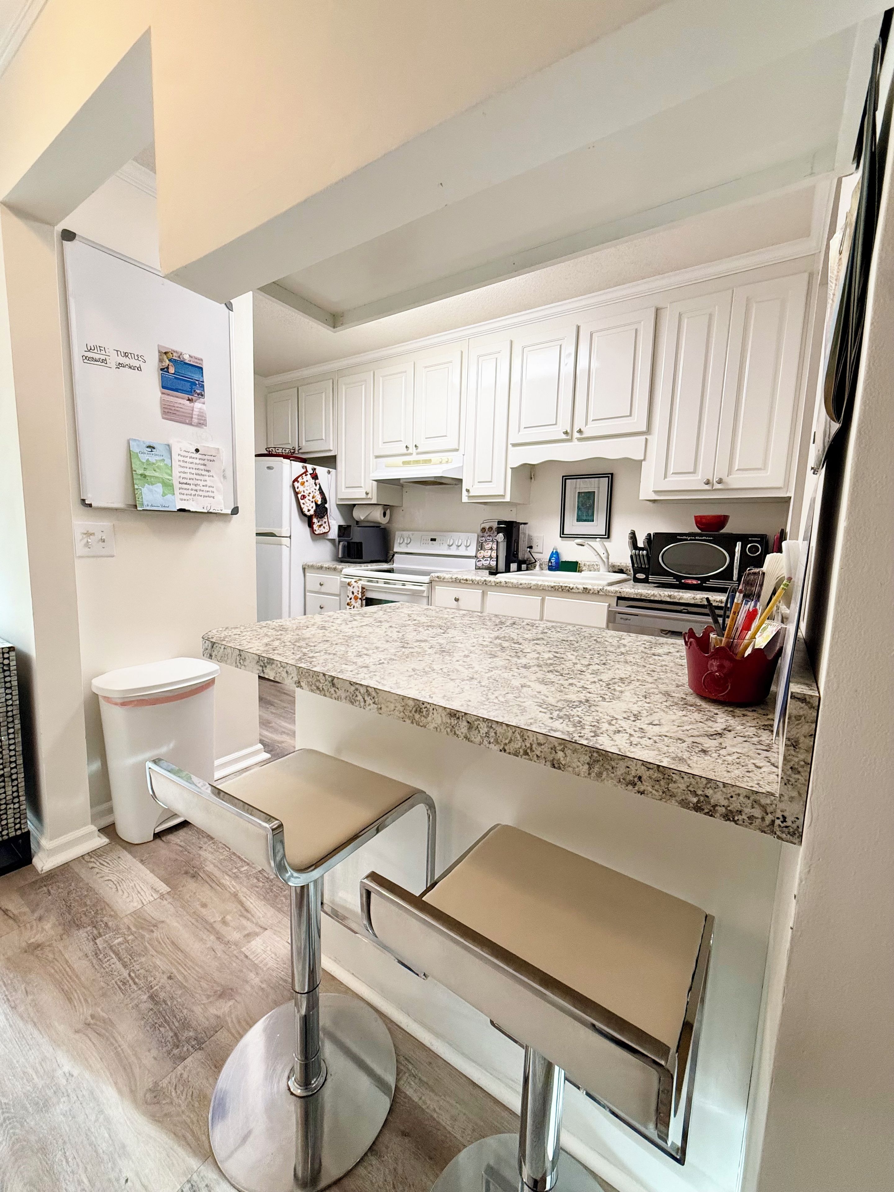 A kitchen with white cabinets, a granite countertop, a stove, a microwave, and a sink. There are two bar stools and a white trash can.
