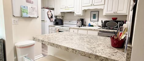 A kitchen with white cabinets, a granite countertop, a stove, a microwave, and a sink. There are two bar stools and a white trash can.