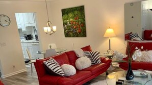Ein modernes Wohnzimmer mit einem roten Ledersofa, einem Glastisch und einem weißen, flauschigen Teppich. Im Hintergrund ist eine Küche erkennbar.