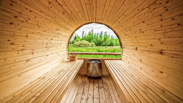 Una sauna de madera con un banco y un recipiente metálico, rodeada de paredes de madera y una ventana con vistas a la vegetación.