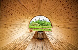 A wooden sauna with a bench and a metal container, surrounded by wooden walls and a window offering a view of greenery.