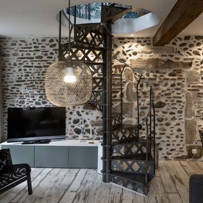 A modern living room with a stone wall, a black spiral staircase, a woven light fixture, a flat-screen TV, and a wooden floor. - A quiet house in the historic district of Pau   (Pau)