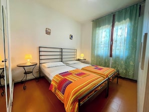 A single-bed room with a metal headboard, two bedside tables with lamps, a colorful striped bedspread, and a green curtain with a floral pattern.