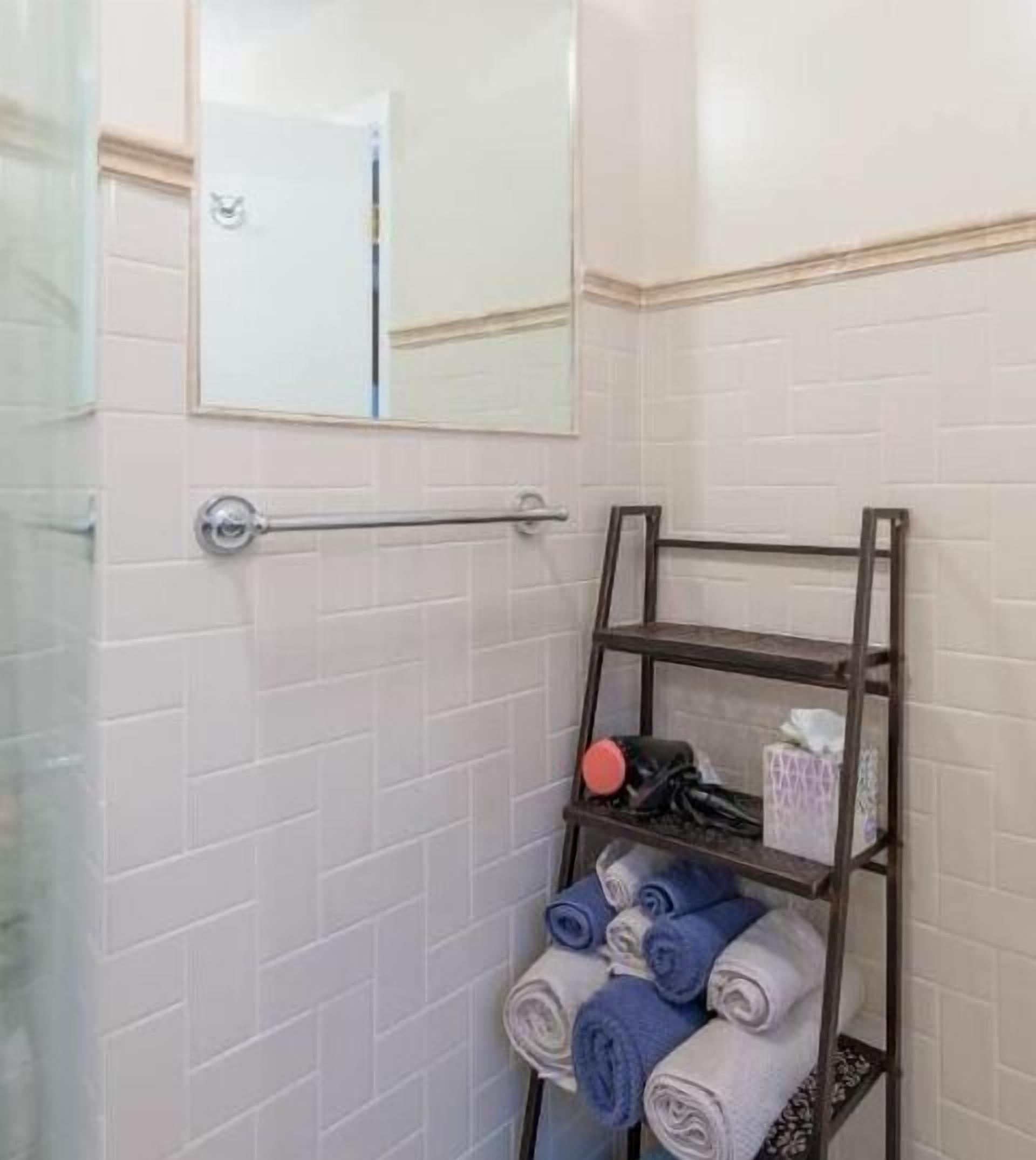 Une salle de bain avec des murs carrelés de carreaux blancs, une douche en verre, un porte-serviettes avec des serviettes pliées et une étagère contenant des articles de toilette.