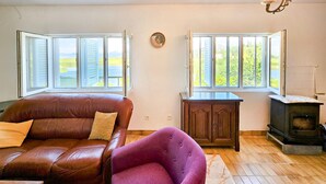 Een woonkamer met een bruine leren bank, een paarse fauteuil, een houten kast, een kachel en grote ramen met uitzicht op groen.