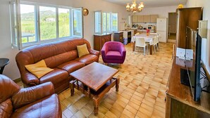 Een woonkamer met een bank, fauteuil en een tafel, een keuken met een eettafel, en uitzicht op groen buiten.