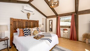A bedroom with a wooden headboard, a bed with white bedding, a nightstand with a lamp, a mirror, and a window with curtains. - The Carson at Ridgeview. Asheville rental! Luxury Glamping! (Alexander)