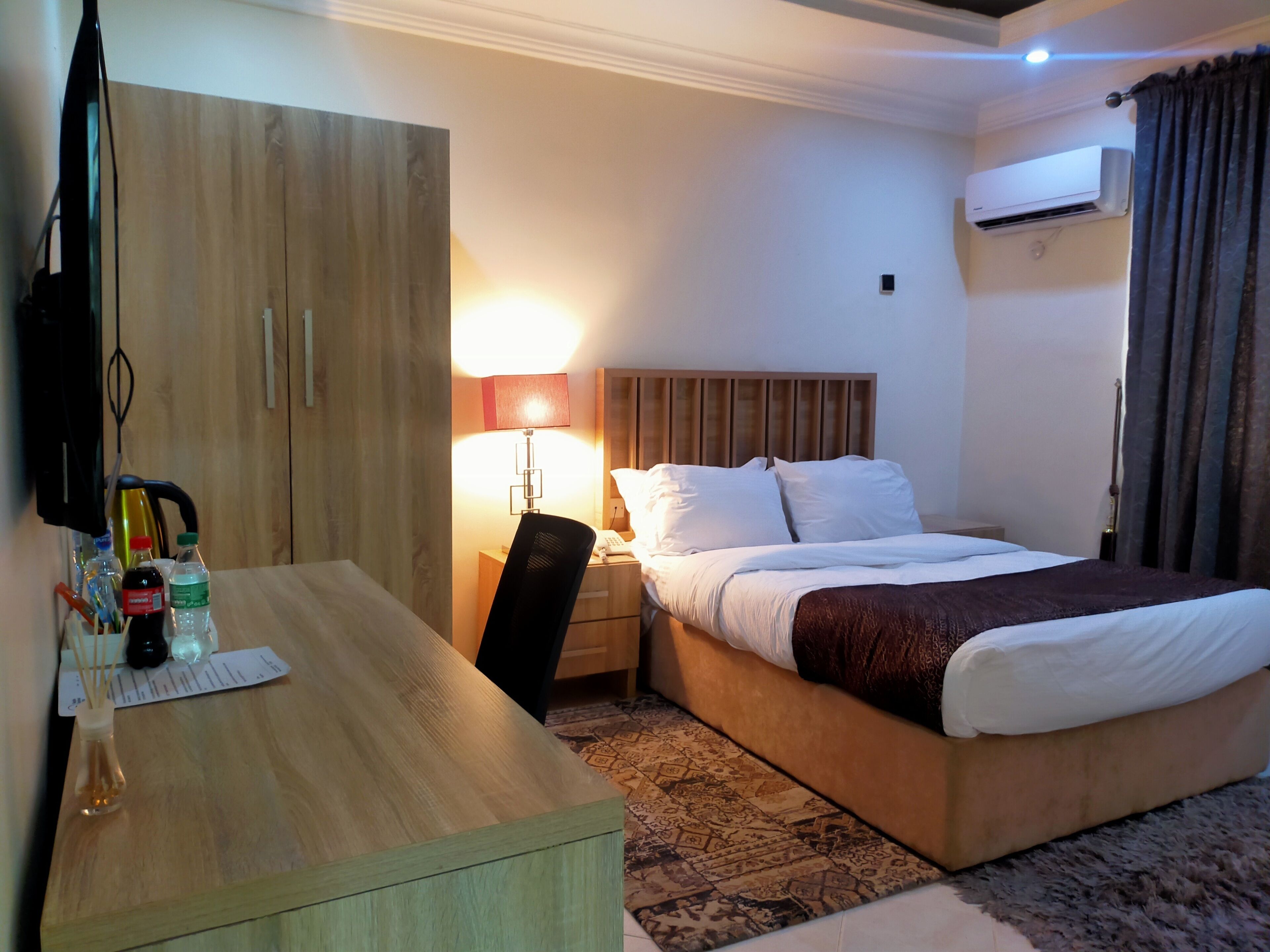 A hotel room with a bed, a desk, a chair, a lamp, and an air conditioning unit.