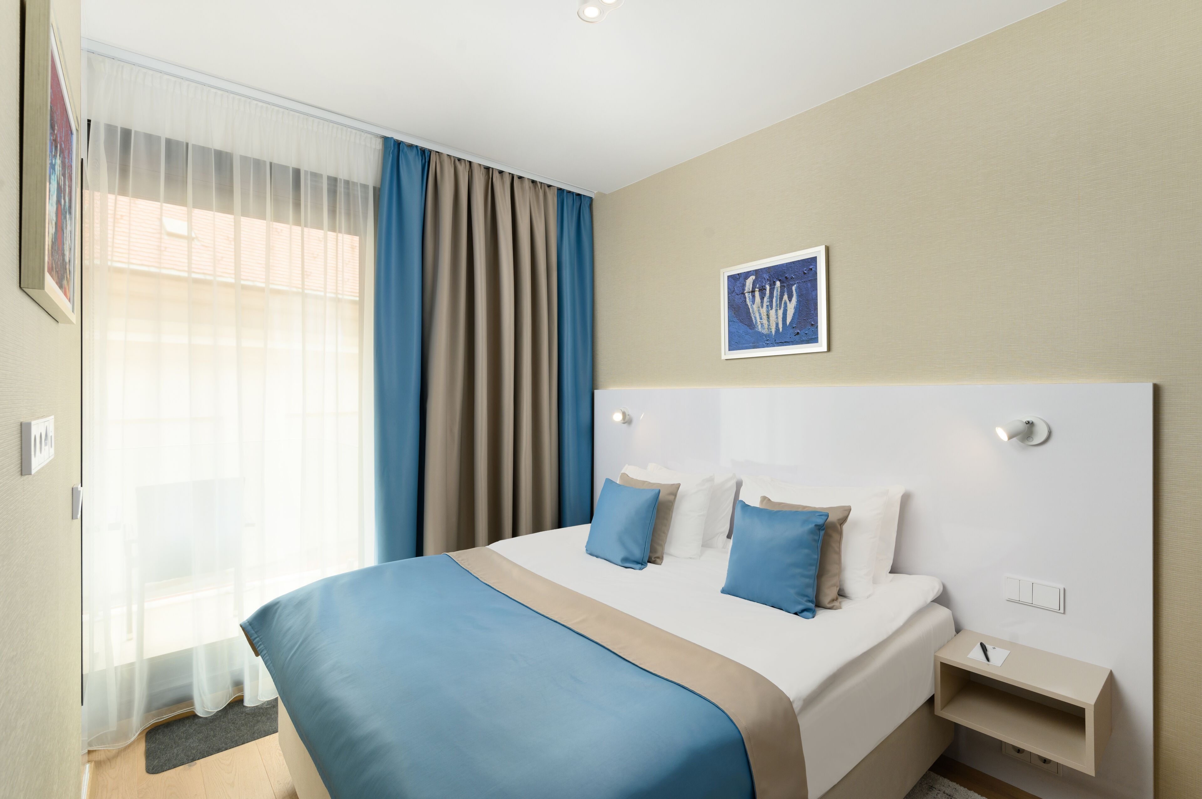 Ein Hotelzimmer mit einem großen Bett, einem Nachttisch, einem Fenster mit transparenten Vorhängen, einer blau-beigen Tagesdecke und einem gerahmten Bild an der Wand.