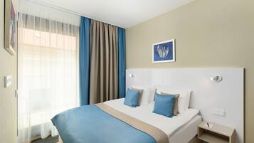 Ein Hotelzimmer mit einem großen Bett, einem Nachttisch, einem Fenster mit transparenten Vorhängen, einer blau-beigen Tagesdecke und einem gerahmten Bild an der Wand.