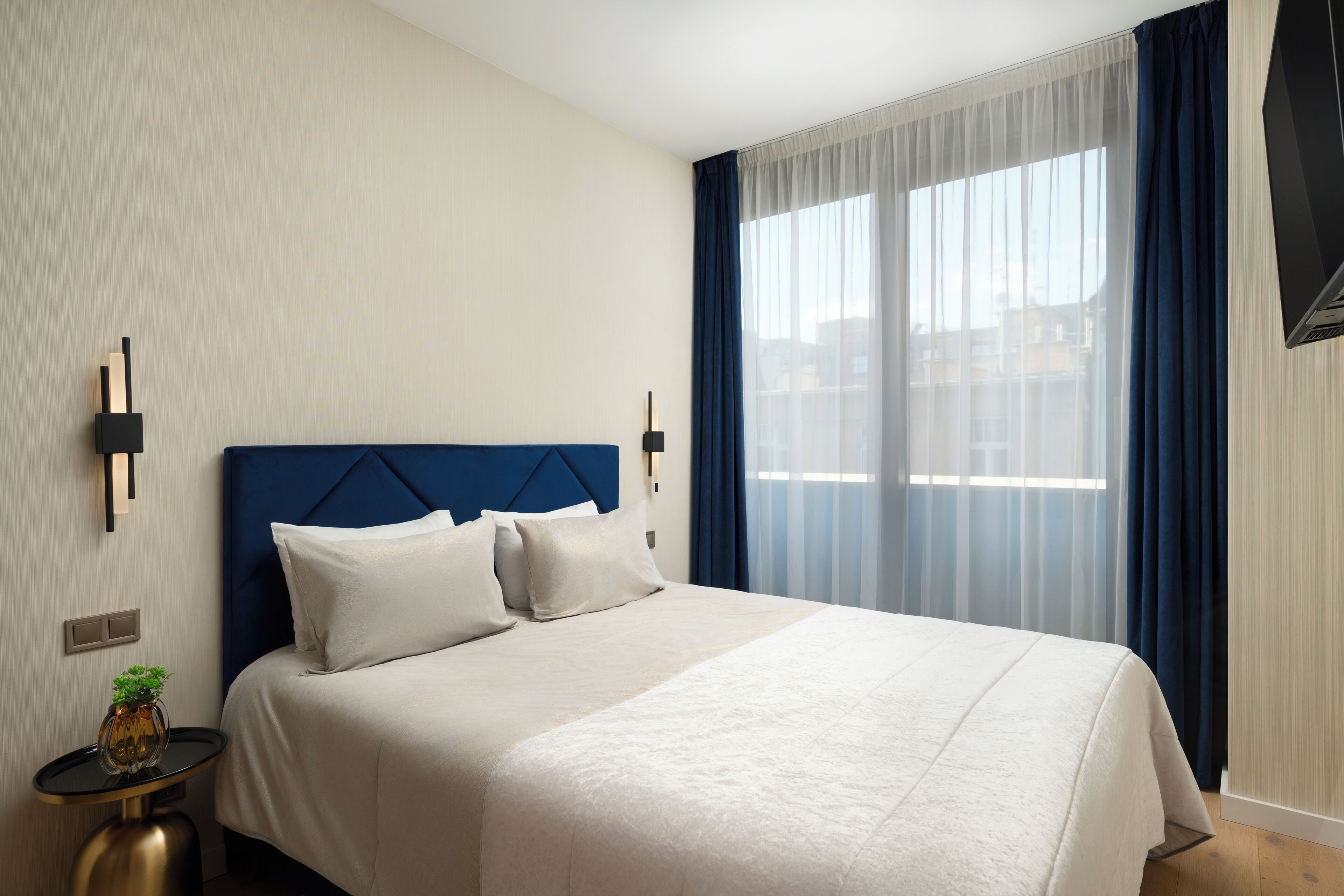 A hotel room with a bed, a side table with a plant, a wall-mounted TV, and a large window with curtains.