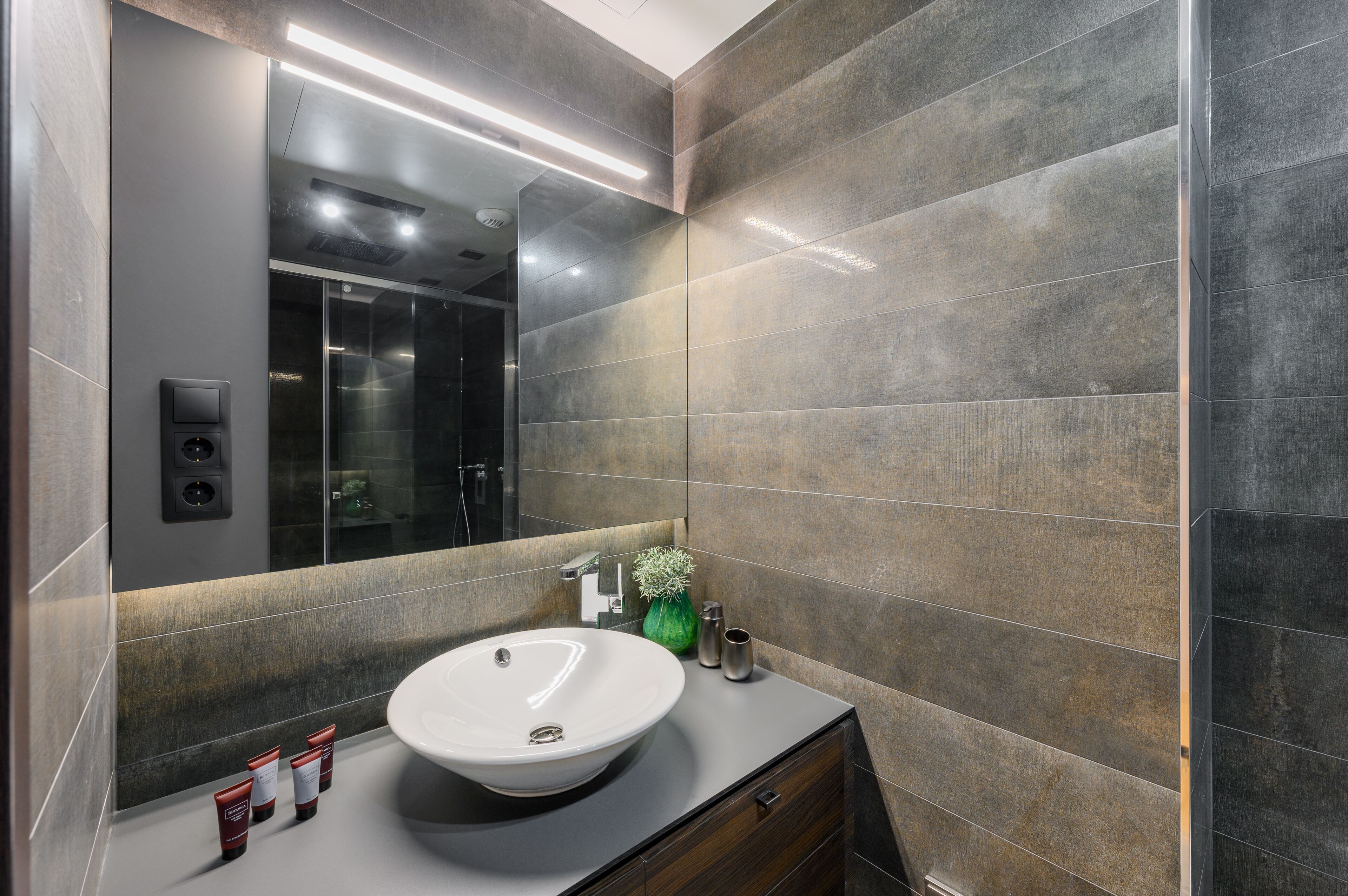 Un bagno moderno con un grande specchio, un lavandino rotondo e una parete rivestita di piastrelle scure.