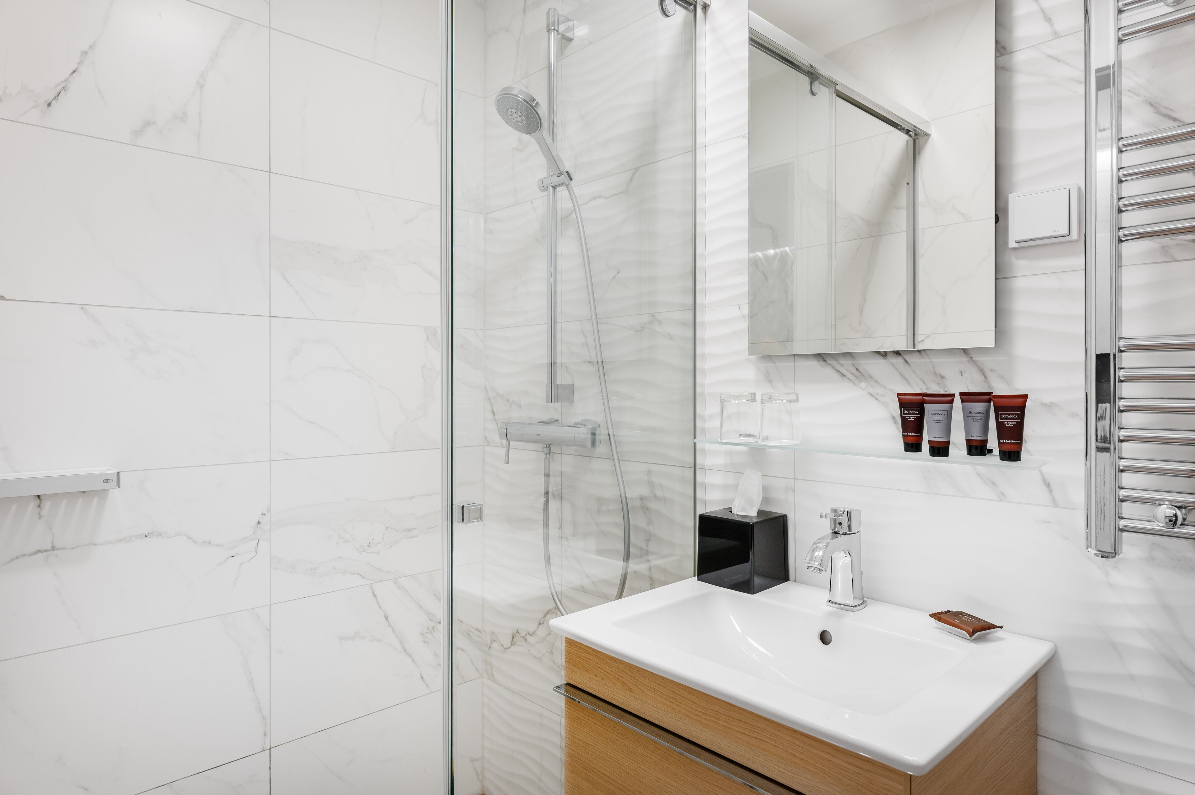 Une salle de bain moderne avec une douche à parois de verre, un mur recouvert de carreaux de marbre blanc, un meuble-lavabo en bois avec un lavabo blanc et un miroir au-dessus du meuble à miroir.