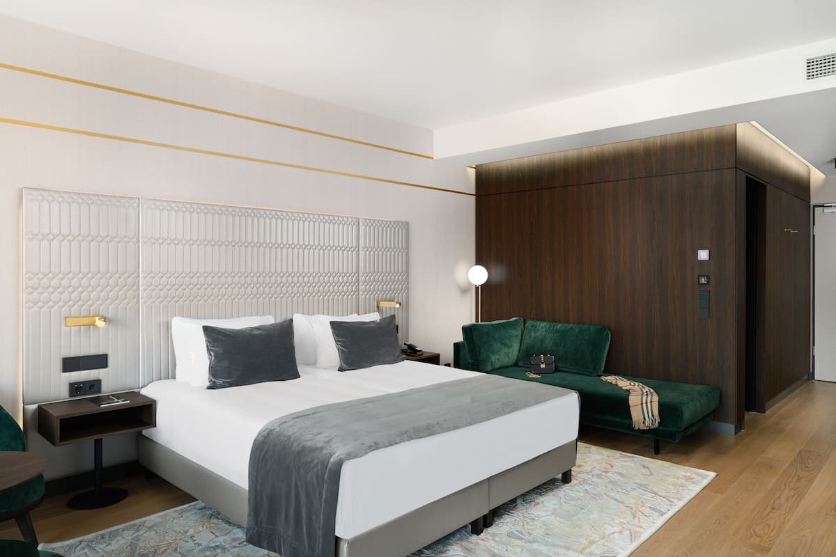 A modern hotel room with a large bed, a green armchair, a wooden wardrobe, and a nightstand with a lamp.