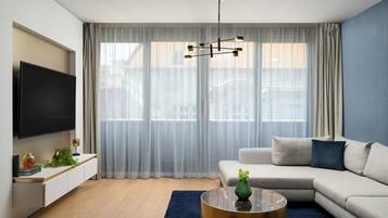 A modern living room with a flat-screen TV, a sofa, a coffee table, and a large window with sheer curtains.