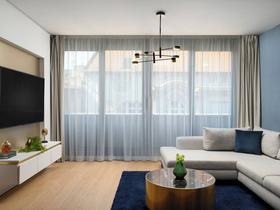 A modern living room with a flat-screen TV, a sofa, a coffee table, and a large window with sheer curtains.