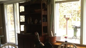 A dining area with a wooden table, chairs, a rooster centerpiece, a bookshelf, and a window with a view of trees.