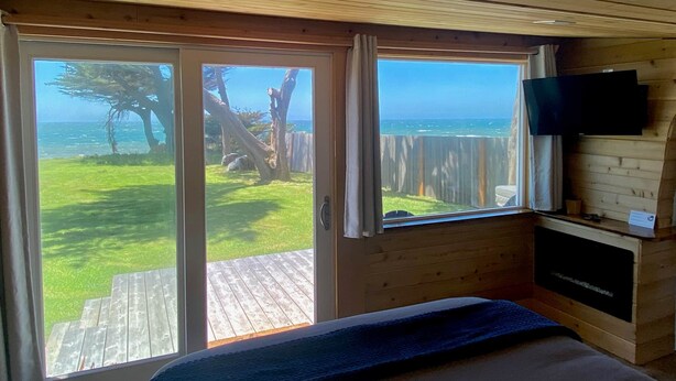 Un dormitorio con techo de madera, una cama, un televisor montado en la pared y un ventanal corredero que da a una terraza con vistas al océano y árboles.