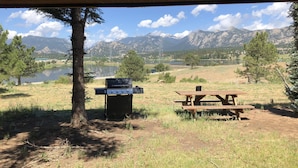Un coin pique-nique aménagé avec un barbecue et une table, dans un cadre naturel avec des montagnes en arrière-plan.