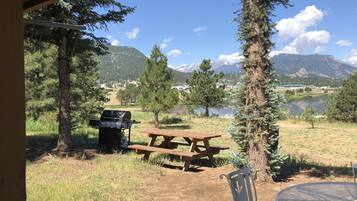 A picnic setup with a grill, table, and chairs in a natural setting with mountains and a lake in the background.