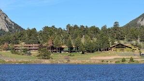 Une propriété située au bord d’un lac, comprenant plusieurs bâtiments, dont un chalet principal et des cabanes, le tout entouré d’une forêt dense et de montagnes.