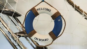 Un bozzello con la scritta "WELCOME ONBOARD", legato a una corda, sullo sfondo di un muro bianco con una ringhiera metallica.