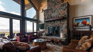 A cozy living room with a stone fireplace, leather furniture, and a large window offering a view of the mountains. - Amazing 3-level home.  Perfect for 3-generation family visits.  Sunsets & elk. (Angel Fire Resort)
