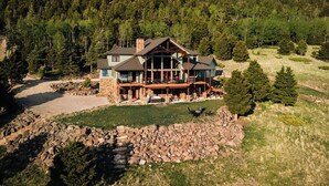 A large, multi-story lodge with a stone facade and a wrap-around deck, surrounded by a forested landscape. - Amazing 3-level home.  Perfect for 3-generation family visits.  Sunsets & elk. (Angel Fire Resort)