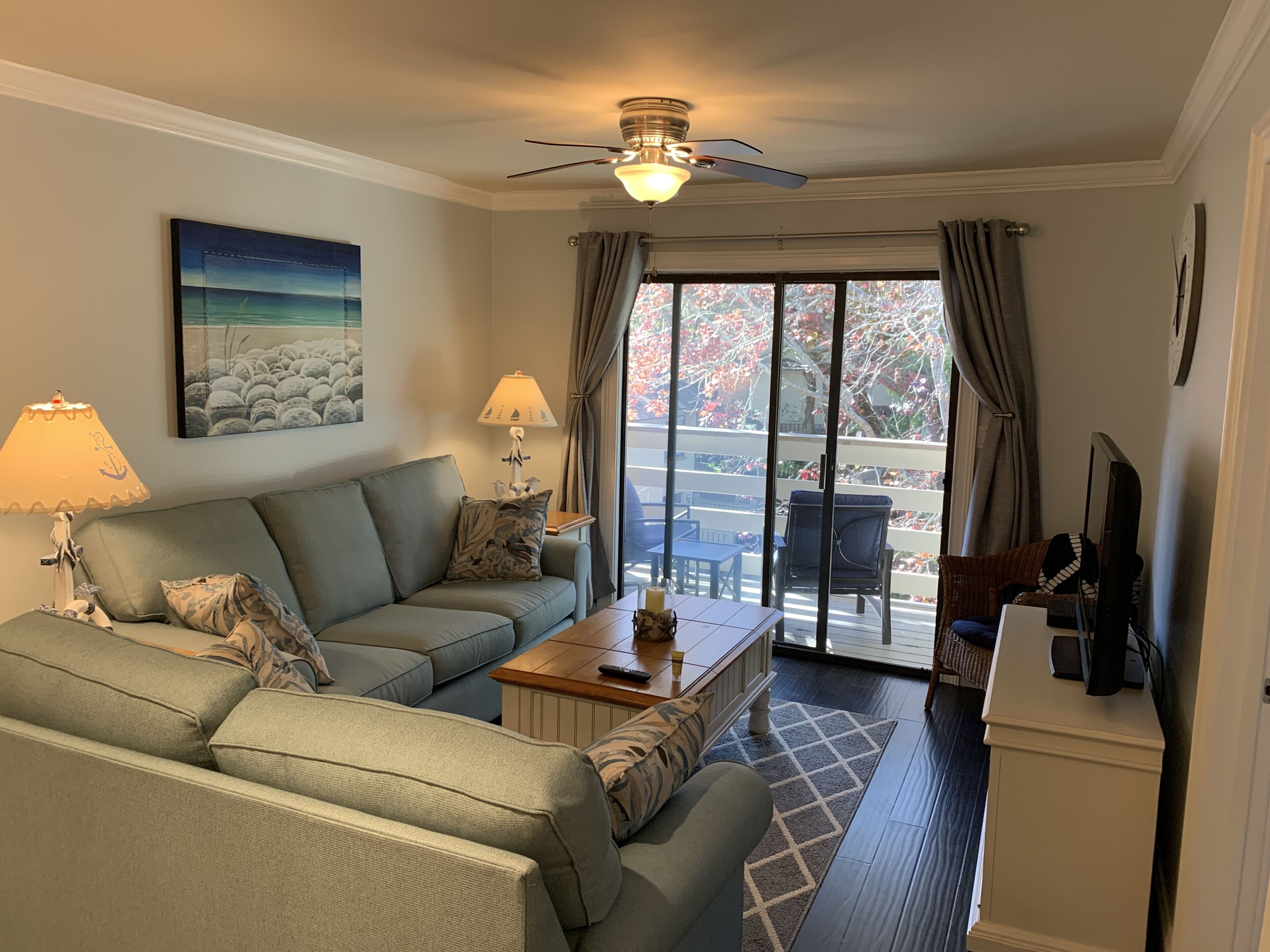 A living room with a sofa, a coffee table, a TV, and a balcony with outdoor furniture.