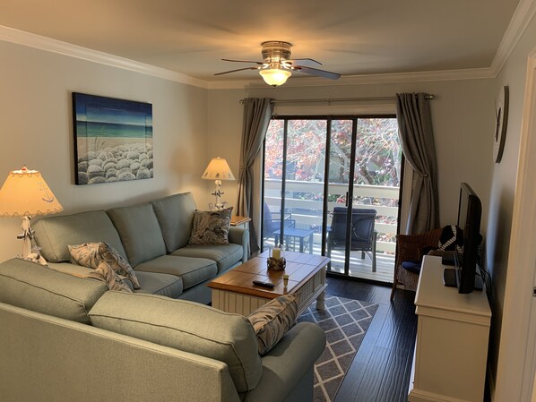 Una sala de estar con sofá, una mesa de centro, televisor y un balcón con mobiliario exterior.