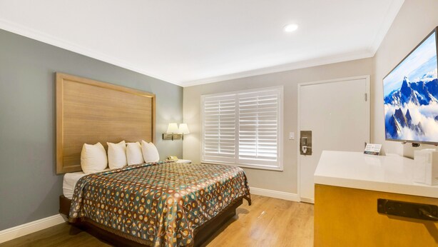 A bedroom with a bed, bedside lamps, a window with blinds, a television, and a wooden cabinet. - Hotel 414 Anaheim, Luxurious King Size Bed (Anaheim)