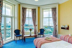 A hotel room with a large bay window, two chairs, a small table, a bed, and a view of a cityscape.