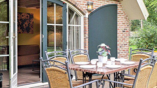 Une terrasse avec une table dressée pour quatre personnes, entourée de chaises en osier, et une porte vitrée donnant sur un espace intérieur.