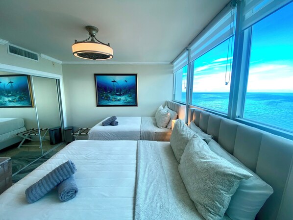 A hotel room with two beds, a ceiling fan, a painting of an underwater scene, and a large window with a view of the ocean.