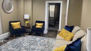 Ein Wohnzimmer mit zwei dunkelblauen Sesseln und goldenen Kissen, einem beigen Sofa mit blauen und goldenen Zierkissen, einem gemusterten Teppich und einer Wanddekoration.