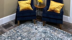 Ein Eckzimmer mit zwei blauen Sesseln, einem kleinen runden Tisch mit einer Lampe und gelben Blumen, sowie einem gemusterten Teppich.