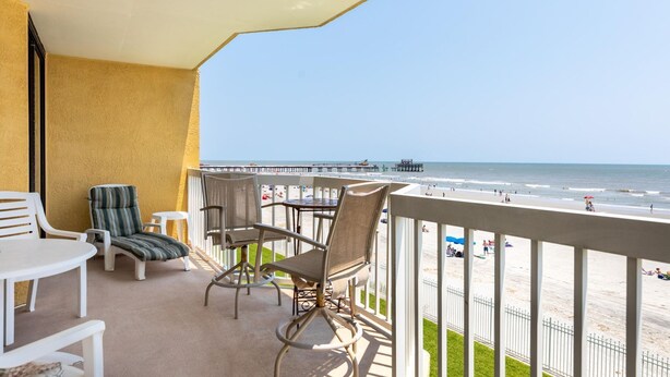 A balcony with a view of the beach, featuring a table and chairs, and a railing.