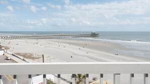 A beach with a long pier extending into the ocean, a clear sky, and a few people on the sand.