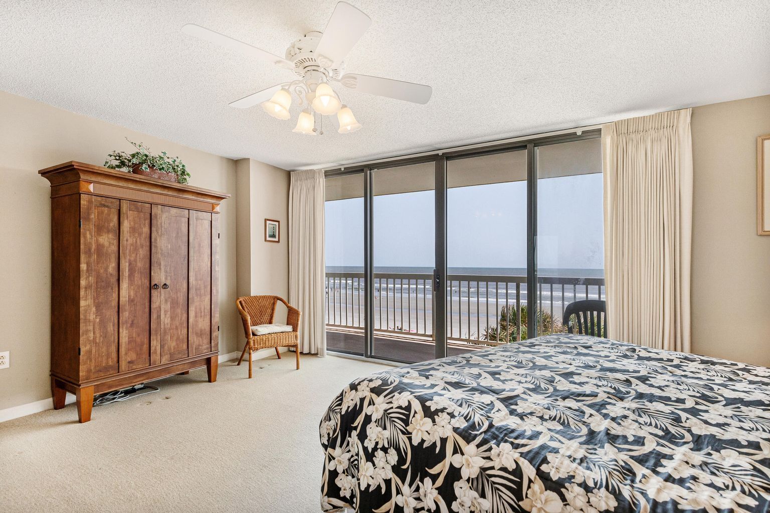 Ein Schlafzimmer mit einem großen Bett, einem Holzschrank, einem Korbstuhl und einem Balkon mit Blick auf den Strand.