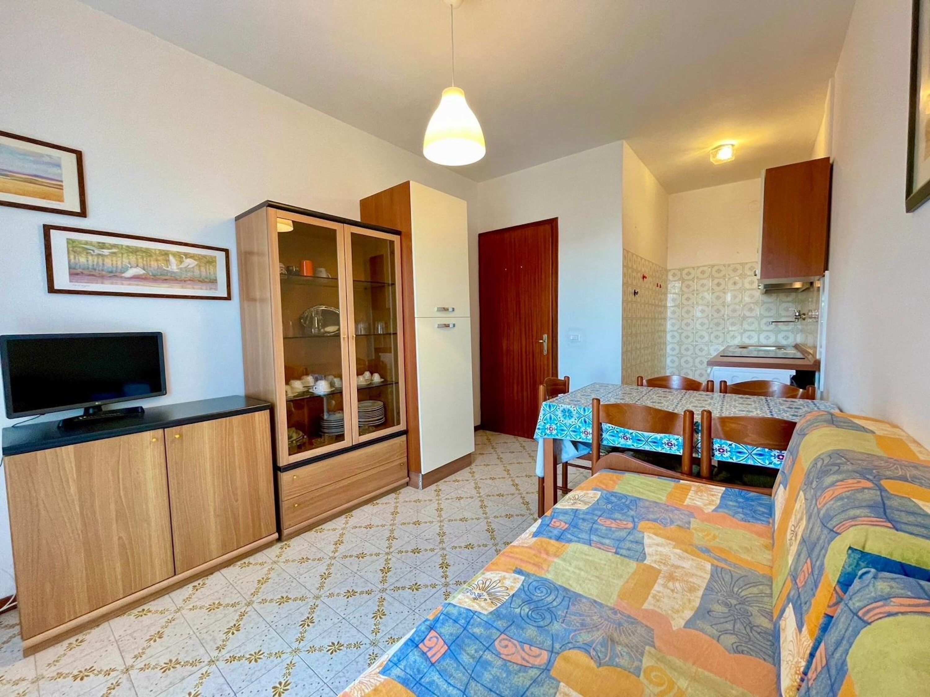 A compact living space with a television, a cabinet with glass doors, a dining table with chairs, and a kitchen area.