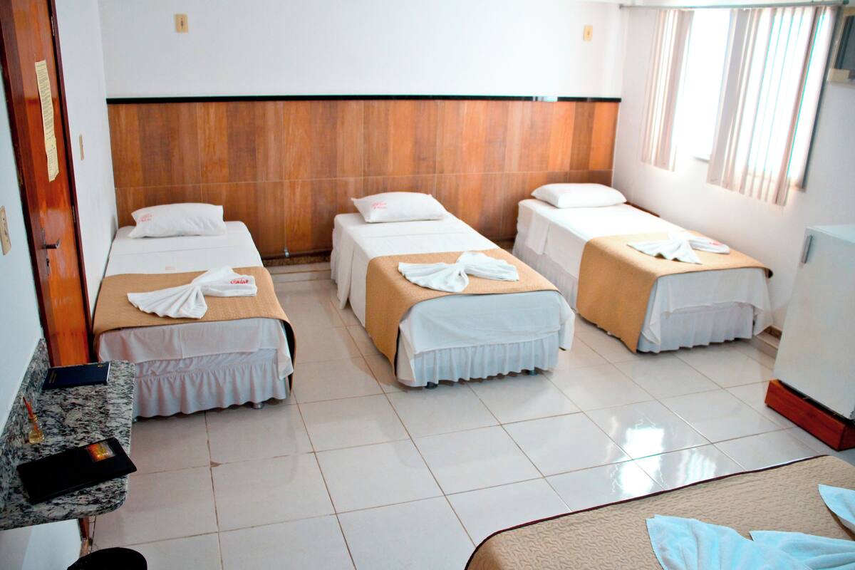 A hotel room with four single beds, each with a white bedspread and a towel. There is a wooden headboard, a window with curtains, and a small table with a book and a bottle.