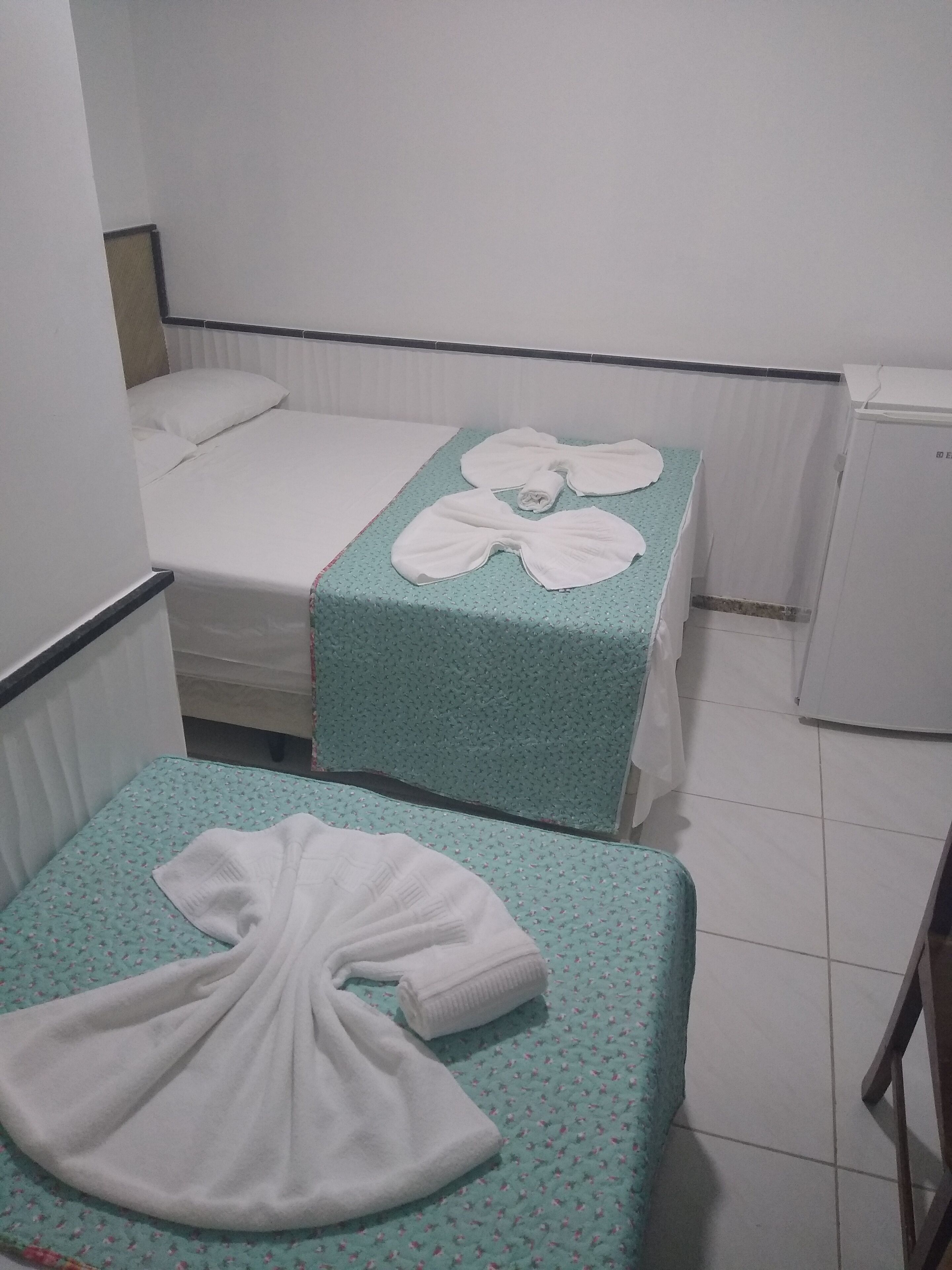 Quarto de hotel pequeno com duas camas, cada uma com colcha azul-turquesa e travesseiros brancos. Há uma pequena mesa com toalha azul-turquesa e uma toalha branca dobrada.