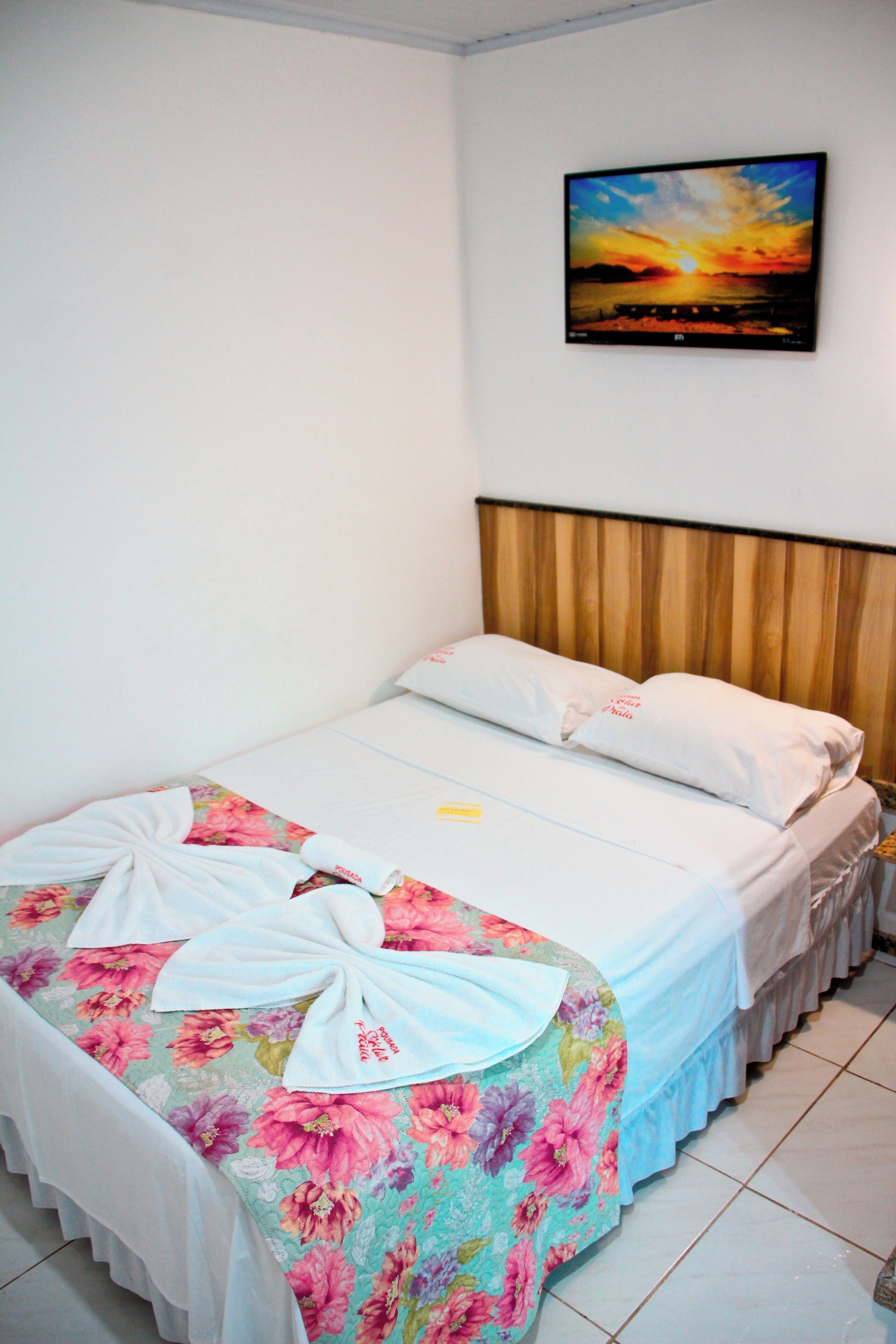 A neatly made bed with floral bedding, a wooden headboard, and a painting of a sunset on the wall.